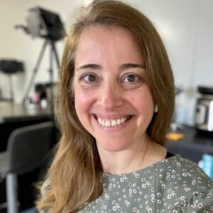 A woman with brown hair smiles for the camera, she wears a green top with a ditsy flower print