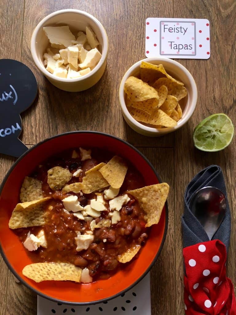 Photo of the Feisty Tapas Mexican Bean soup close-up with ingredients to serve. Soup is served in an orange bowl and smaller pots hold the tortillas / Doritos and the cheese, there's a wedge of lime and a grey napkin with a red and white polka dot ribbon holding cutlery. There's also a blackboard with the name of the recipe, Feisty Tapas Mexican Bean Soup