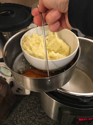 Photo of the Kuhn Rikon steamer basket showing being held above an Instant Pot by a hand, in the basket there is a bowl with mash and a few carrot batons