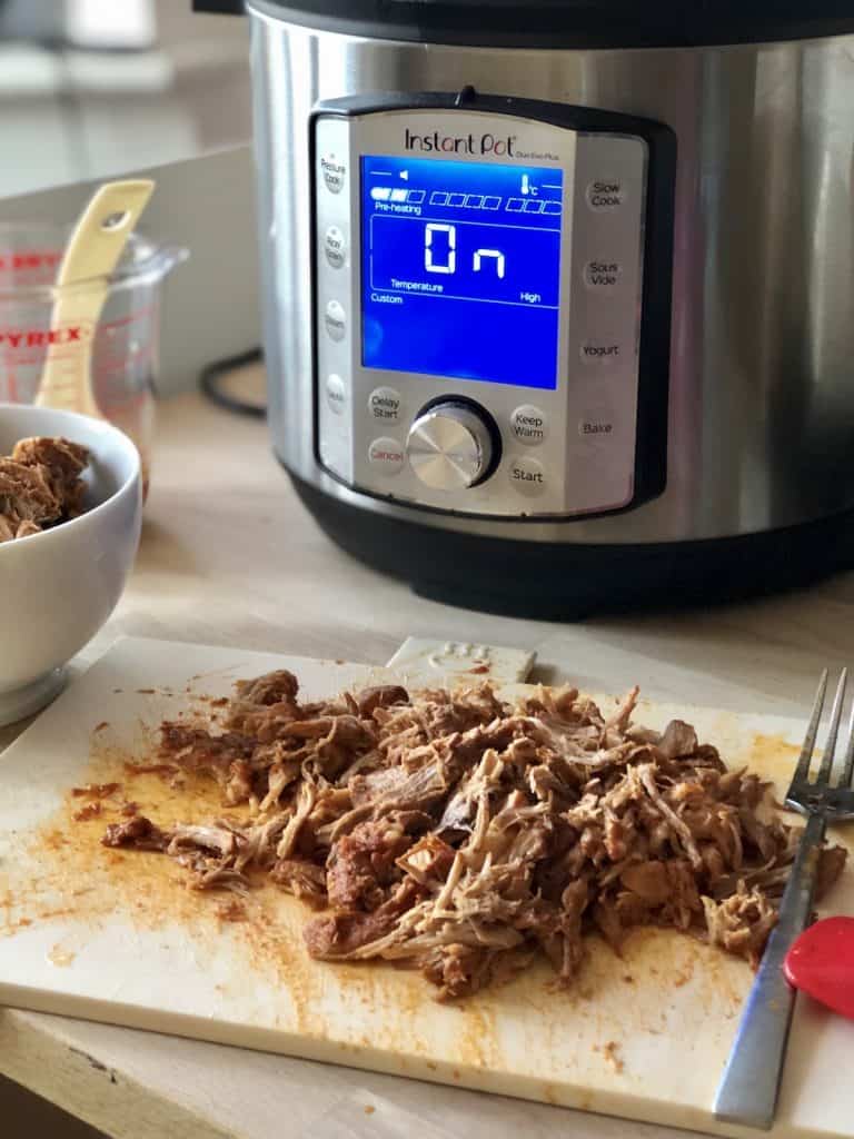 The image shows the pressure cooked pulled pork, there's an Instant Pot in the background, a fork on the right and white bowl