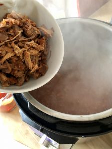 Image shows a bowl of pulled pork being held above an Instant Pot from which a lot of steam is rising