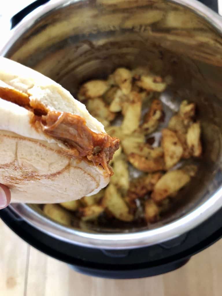 Photo of a bun filled with Feisty Tapas Easy BBQ Pulled Pork being held over a pot full of Greek Roast Potatoes