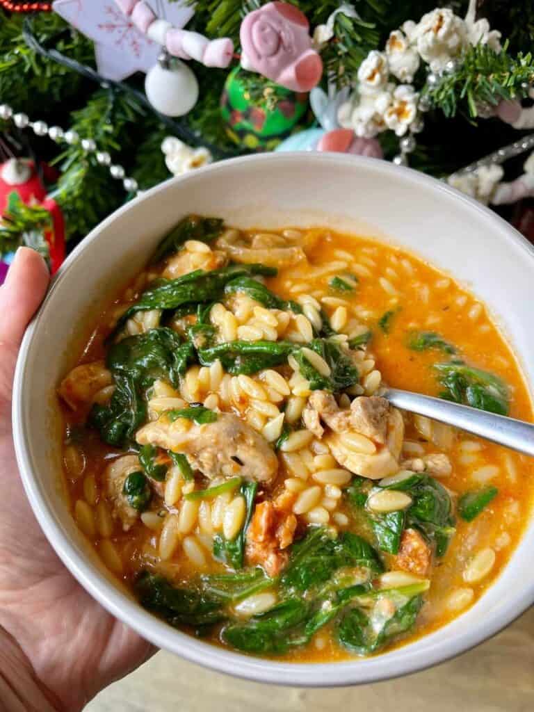 Chicken and Chorizo Orzo Pasta recipe served in a white bowl with a Christmas tree in the background. Shown here held by a left hand