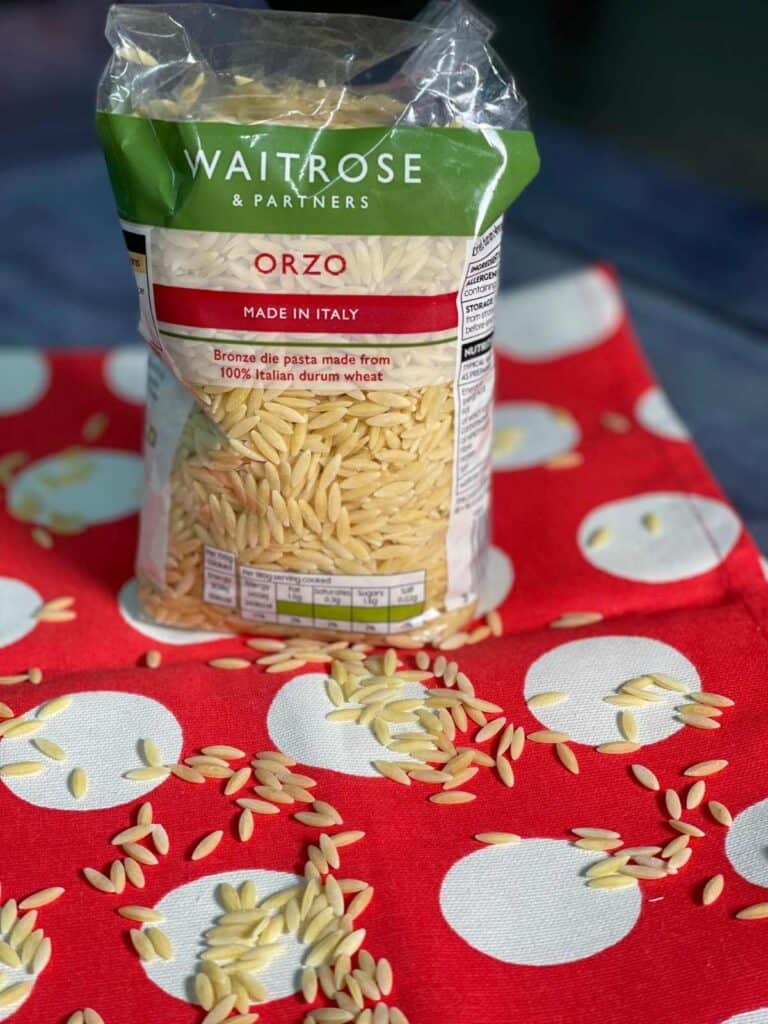 A bag of Waitrose Orzo pasta standing on a red and white polka dot fabric, with rice-shaped orzo risoni pasta on the fabric too