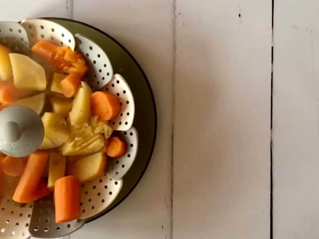 Carrot and swede chunks seen from above in a Joseph Joseph white and grey steamer basket, on a white surface