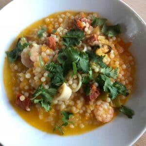 Photo of the served giant cous cous dish seen on a deep white plate, with the vibrant broth, squid rings, tiger prawns, chorizo and mussels on show and finished with fresh coriander