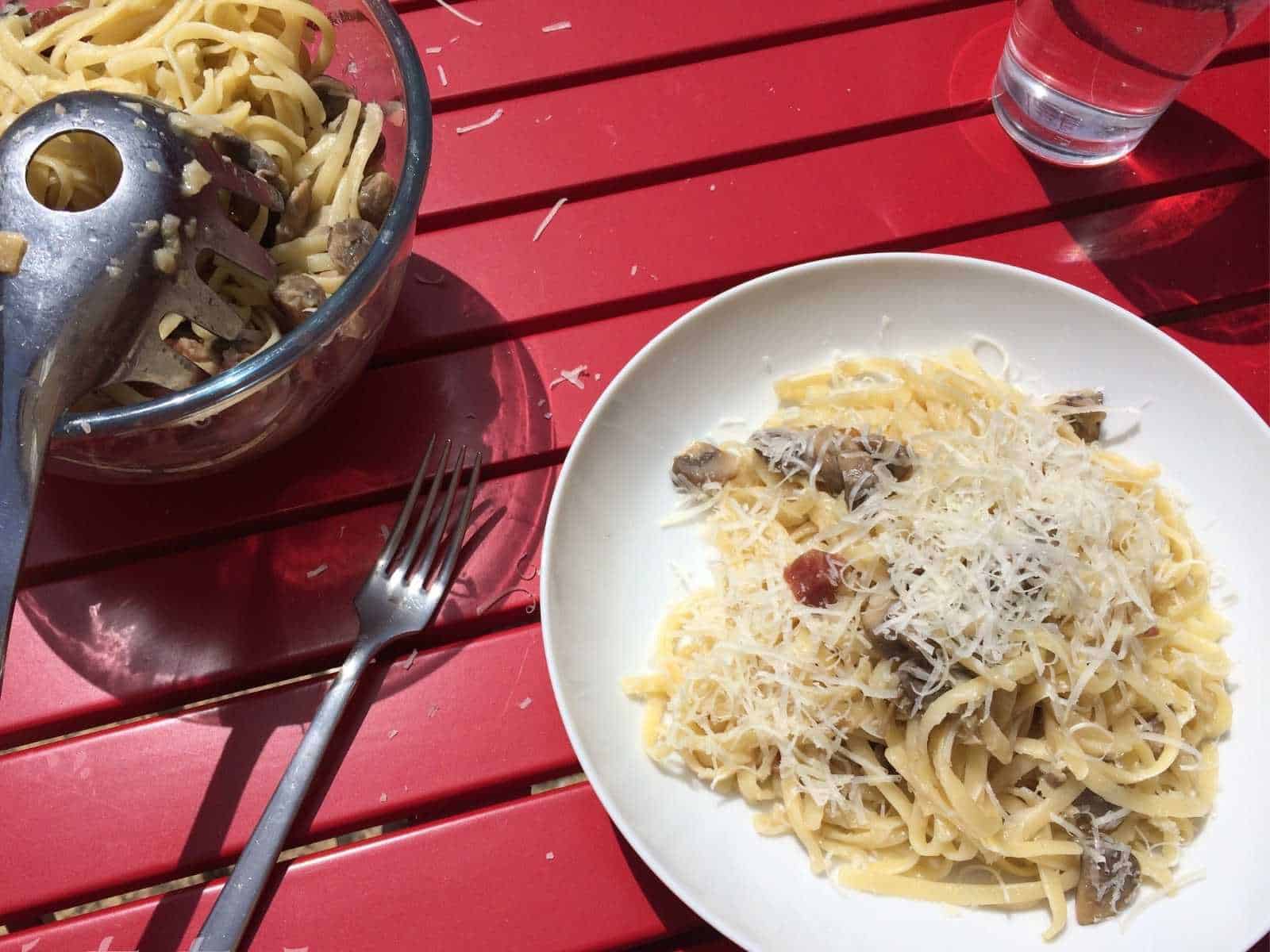 Thermomix Garlic and Serrano Ham Mushrooms (Spanish Setas al ajillo) served with linguine and topped with parmesan cheese. Seen against a red slatted table with a pyrex bowl full of mushrooms on the left
