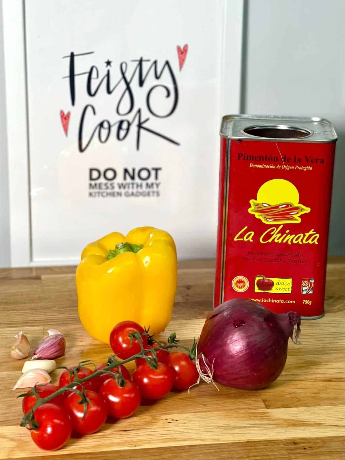 Ingredients for Air Fryer roasted vegetables laid on a wooden chopping board. Cherry tomatoes on the vine, red onion, yellow pepper, garlic cloves and a giant La Chinata smoked paprika metallic tub