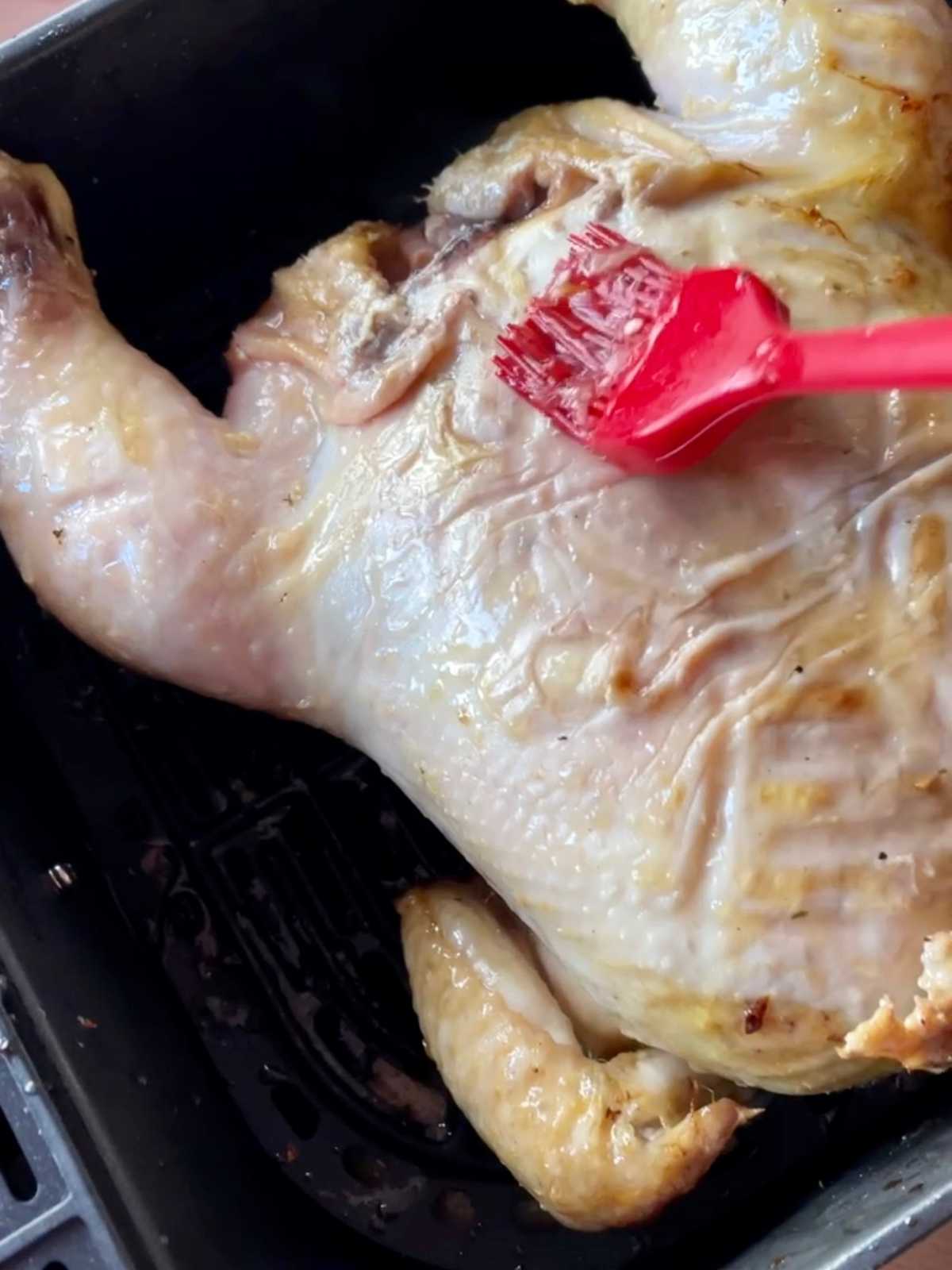 Chicken seen from above, breast-side now up with a red silicone brush brushing the glaze to make it juicy