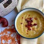 Pressure Cooker Chorizo and Red Lentil Soup seen from above inside a blue bowl with white inside. The bowl is resting on a yellow linen napkin. There are red lentils scattered on the white surface on the left, a chorizo sausage hoop with a chunk missing and an Instant Pot Duo Crisp