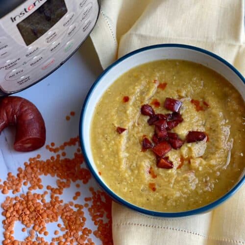 Pressure Cooker Chorizo and Red Lentil Soup seen from above inside a blue bowl with white inside. The bowl is resting on a yellow linen napkin. There are red lentils scattered on the white surface on the left, a chorizo sausage hoop with a chunk missing and an Instant Pot Duo Crisp