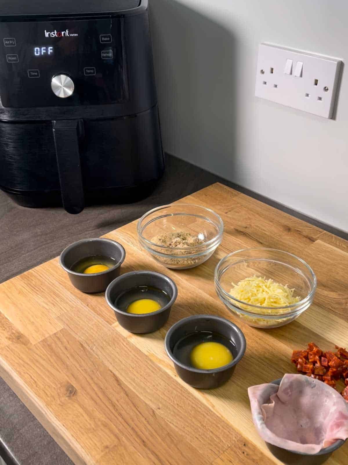 Non-stick mini basins on chopping board with all ingredients laid out and eggs in pots