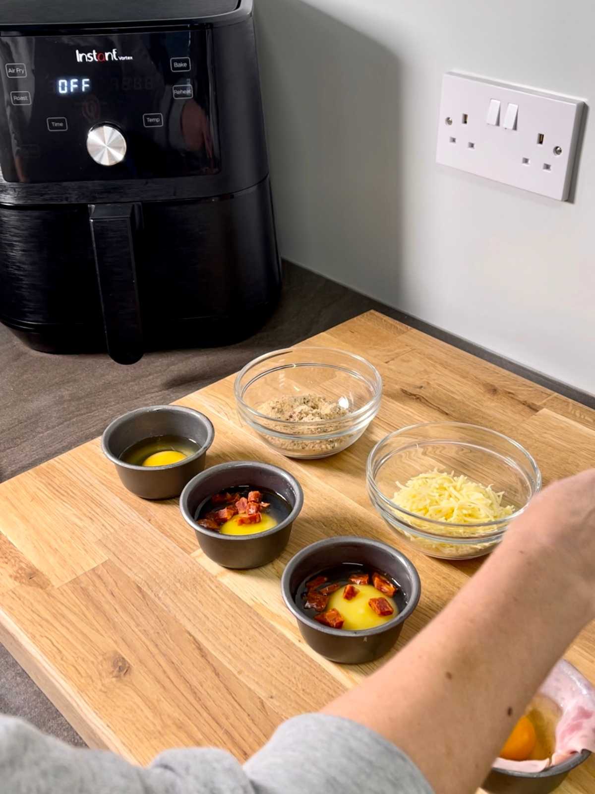 Non stick mini basins on large wooden chopping board with eggs and chorizo inside basins and other ingredients in the shot