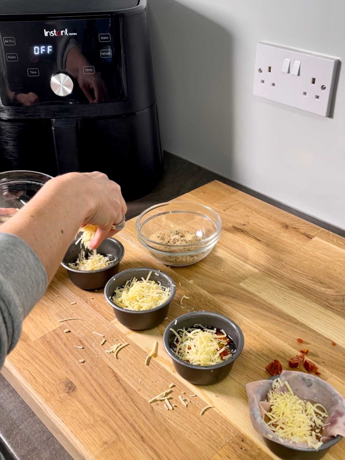 Non-stick pudding basins on large wooden chopping board, a hand is seen added grated cheese