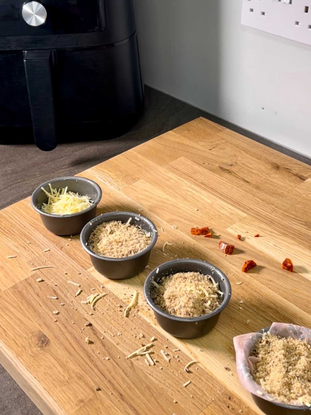 Egg pots on large wooden board, topped with the final ingredient, breadcrumbs, and ready to go in the air fryer