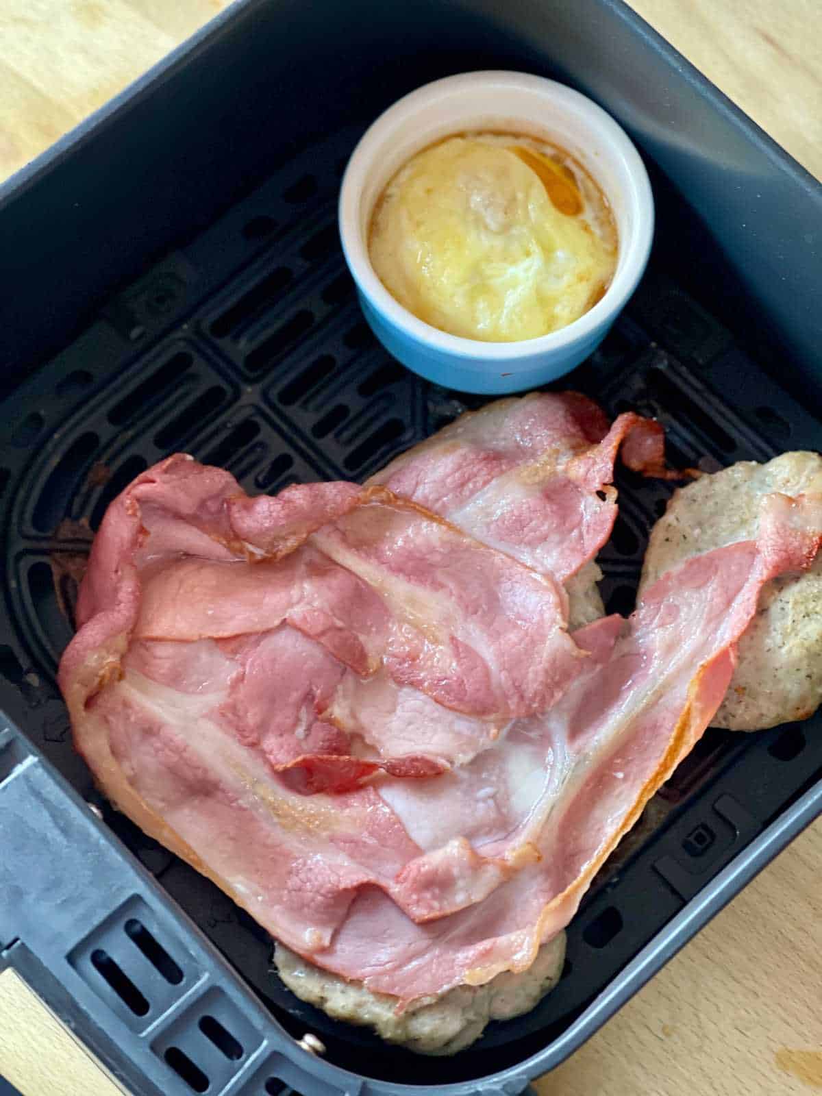 Photo of the bacon on top of the sausage patties half way through cooking and just before flipping over with an egg in a ceramic ramekin with added cheese slices - half way through - Air fryer used is the Instant Vortex