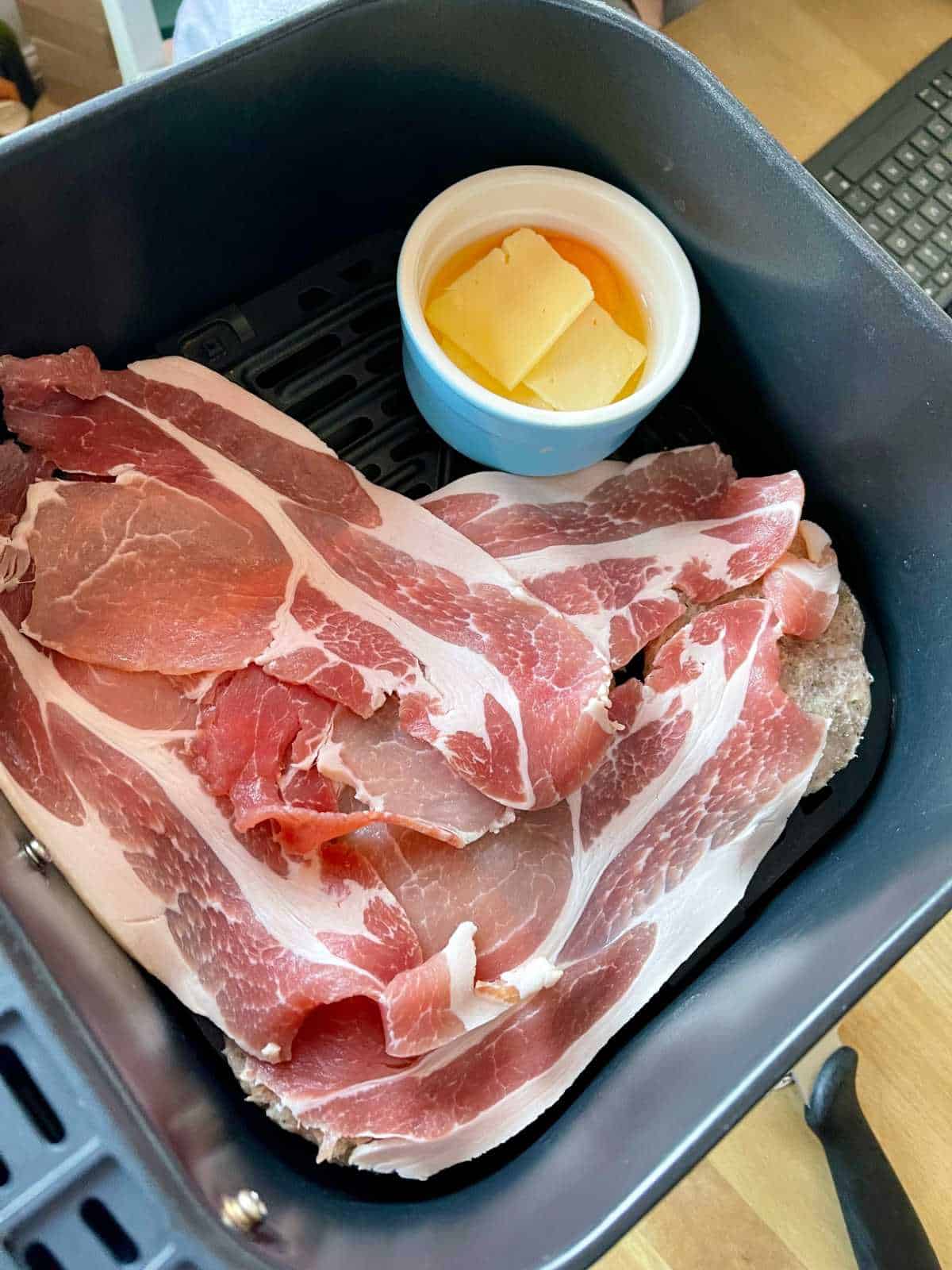 Photo of bacon rashers laid on top of the sausage patties for air fryer McMuffins with a ramekin with an egg and a couple of cheese slices