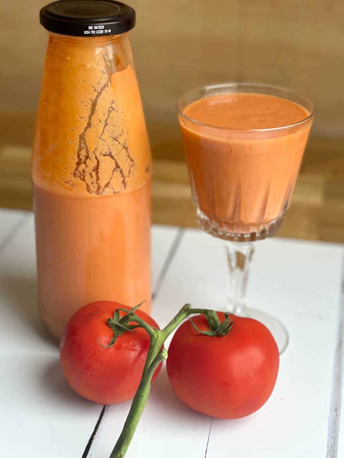 Photo of the final result, on a pretty stem glass on the right there is the chilled Thermomix gazpacho served in a pretty stem glass. On the left there is an old passata bottle half full with gazpacho ready to go in the fridge and in front of these there are two ripe tomatoes on the vine