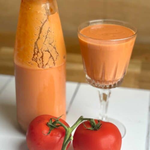 On the right there is a pretty stem glass full of gazpacho, on the left there is a glass bottle half full of gazpacho and in front of them there are two tomatoes on the vine
