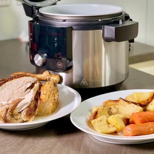 A whole juicy chicken and the vegetables served in two large white plates with the Ninja 14 in 1 in the background. The vegetables include potatoes, carrots and parsnips