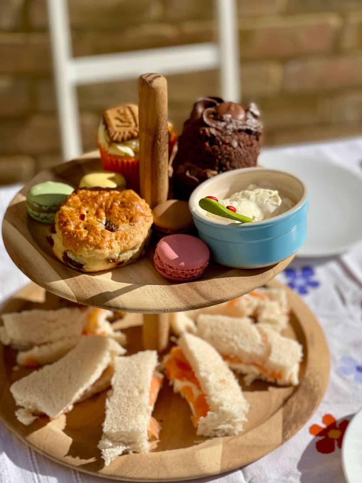 Photo of an afternoon tea served on a two-tier wooden stand, the bottom shelf has smoked salmon and cream cheese sandwiches and the top shelf has macarons, clotted cream, scones and a chocolate cake