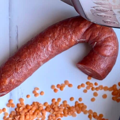 A chorizo sausage loop with a bit cut off seen from above on a white surface surrounded by dried red lentils