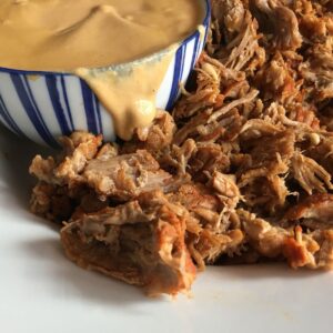 Image of the pressure cooked pulled pork on a white plate with the dairy-free creamy paprika sauce at the back in a stripy white and navy blue bowl