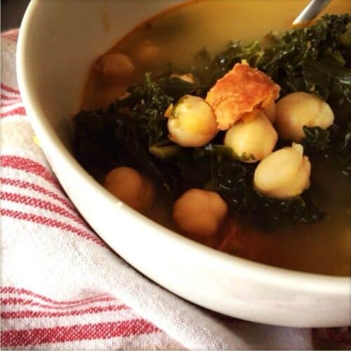 The delicious kale, chickpea and chorizo soup served in a white bowl sitting on a beige tea towel with red stripes