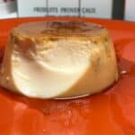 A pressure cooked flan de Huevo served on an orange plate, one bite has already been eaten so the smooth inside is showing, there's delicious caramel too