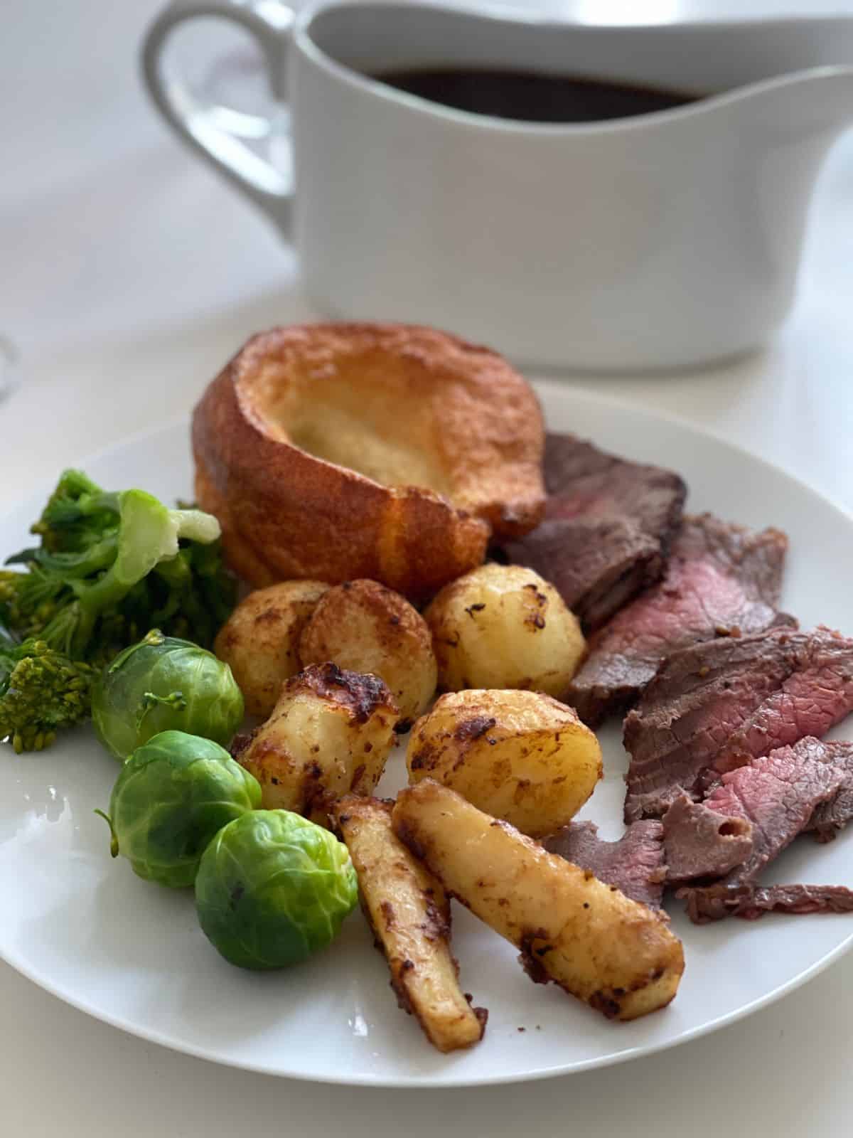 Sliced roast beef, parsnips and potatoes served on a white plate with Brussels sprouts, broccoli and a Yorkshire Pudding