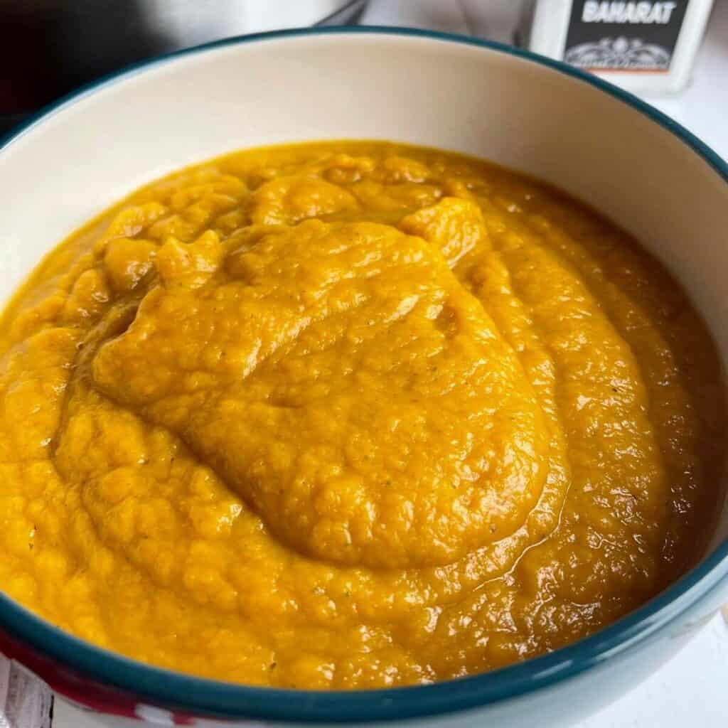 The Roast Pumpkin Soup served in a white bowl which is blue on the outside, there is a small metallic container with the word Baharat on it on the top right