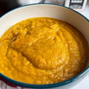 The Roast Pumpkin Soup served in a white bowl which is blue on the outside, there is a small metallic container with the word Baharat on it on the top right