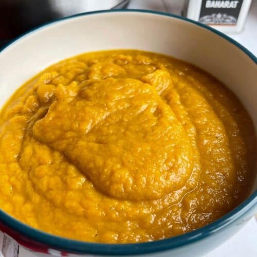 The Roast Pumpkin Soup served in a white bowl which is blue on the outside, there is a small metallic container with the word Baharat on it on the top right