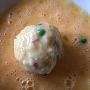 An arancini rice ball coated in beaten egg resting on a plate full of beaten egg