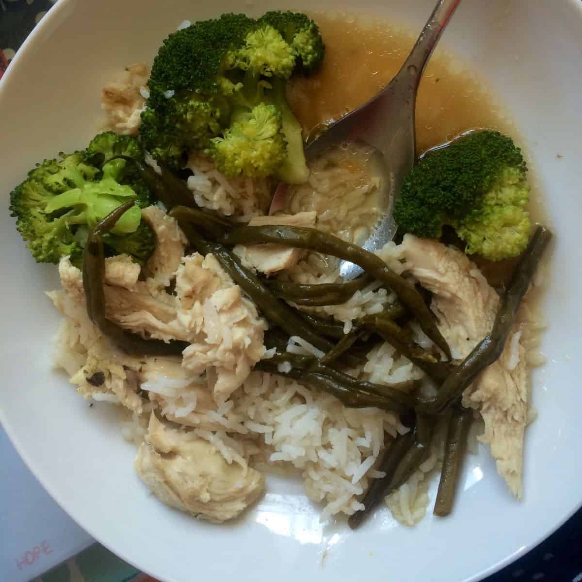 Soy chicken served with rice and steamed broccoli plus the optional overcooked green beans on a plate, there's a spoon an a lot of delicious broth